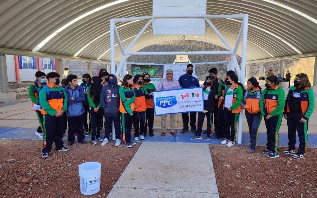 Group of Mexican students in green and orange uniforms standing with PPC team members under a covered basketball court holding a banner thanking Jared Sarbit and Picture Perfect Cleaning.