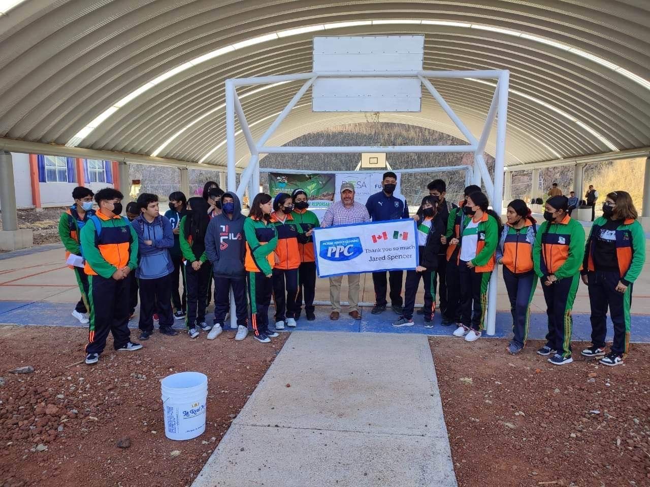 PPC Supporting Environmental Education in Mexico Group of Mexican students in green and orange uniforms standing with PPC team members under a covered basketball court holding a banner thanking Jared Sarbit and Picture Perfect Cleaning.