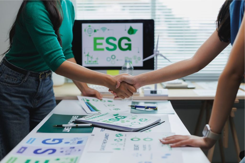 Two team members shake hands over a table filled with ESG compliance reports.