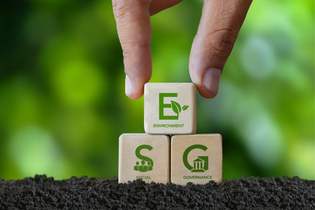 A hand holds three wooden blocks stacked in a pyramid in soil. The top one has a green E and says "Environment," the bottom two, respectively, have an S with the word "Social," and a G, showing the word "Governance."